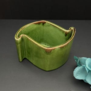 Peut inclure: Un bol en céramique vert avec un design ondulé unique. Le bol a une finition brillante et une bordure marron autour du bord. L'intérieur est de couleur verte unie et l'extérieur a un aspect texturé. Le bol est probablement destiné à un usage décoratif ou fonctionnel.