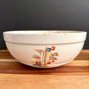 May include: Off-white ceramic mixing bowl with a colorful illustration of a pantry or kitchen scene. The bowl has a wide, rounded shape and a slightly distressed appearance, suggesting vintage charm. The illustration features shelves with various kitchen items.