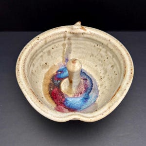 May include: A handmade ceramic apple baker with a central stem for holding the apple. The bowl is beige with brown speckles and a colorful swirl of red, blue, and brown glaze at the base of the stem. The baker has a small spout.