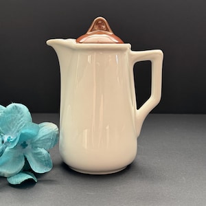 May include: A white ceramic creamer with a brown lid and handle. The creamer has a spout for easy pouring. The lid is a darker shade of brown. The creamer is a classic design, suitable for serving cream or milk.