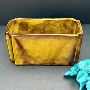 以下が含まれることがあります： 金と茶色の光沢のあるまだら模様の長方形のセラミックプランター。 プランターは、わずかにフレアした側面と濃い茶色の縁を持つ幾何学的な形状をしています。 ターコイズブルーの花が右下隅にあります。