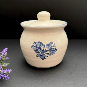 May include: A beige ceramic jar with a lid, featuring blue floral designs. The jar has a rounded shape and a light blue trim on the lid. The background is dark, highlighting the jar's details.