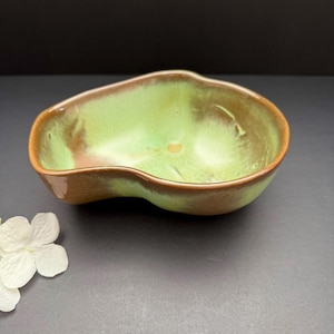 May include: A ceramic bowl with an unusual, organic shape. The interior is a light green, transitioning to a brown rim. The bowl's exterior is a warm brown. A white flower is in the lower left corner.