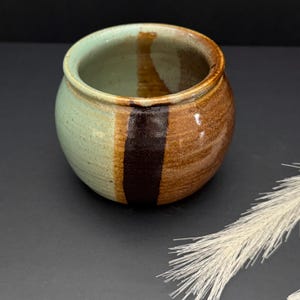 May include: A handmade ceramic pot with a rounded shape. The exterior features a color scheme of light green, brown, and dark brown vertical stripes. The pot has a smooth, glazed finish and a wide opening.