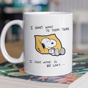 May include: White ceramic mug featuring Snoopy sleeping on a yellow pillow. The mug has the text "I DON'T WANT TO THINK TODAY" and "I JUST WANT TO BE COZY..." printed in black. The mug has a large, curved handle.