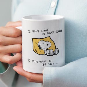 May include: White ceramic mug featuring Snoopy sleeping in a yellow blanket. The mug has the text "I DON'T WANT TO THINK TODAY, I JUST WANT TO BE COZY..." in black lettering. The mug is held by a person.