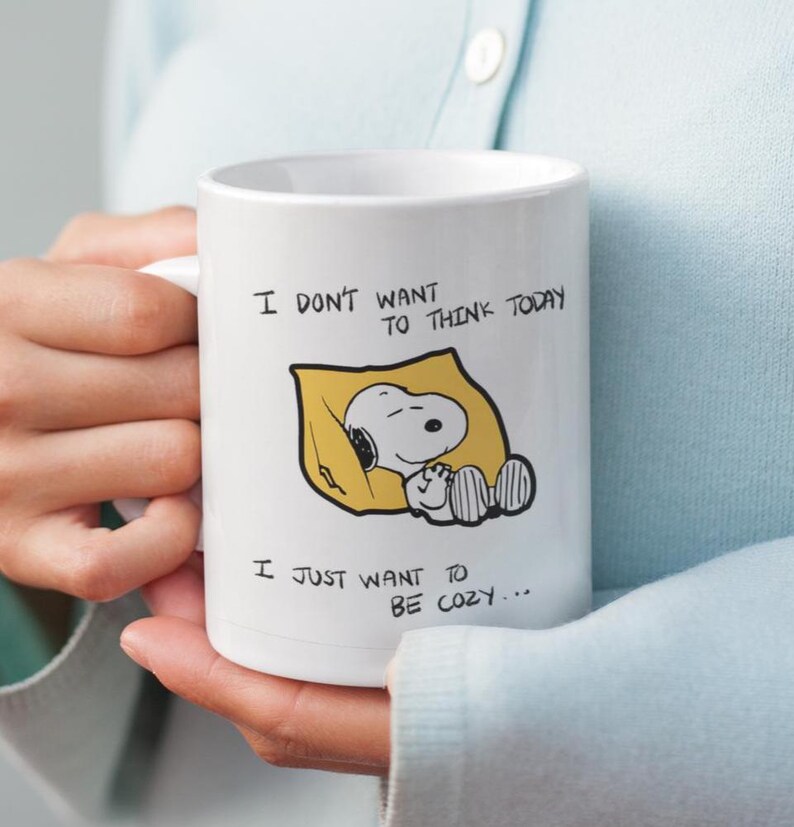 May include: White ceramic mug featuring Snoopy sleeping in a yellow blanket. The mug has the text "I DON'T WANT TO THINK TODAY, I JUST WANT TO BE COZY..." in black lettering. The mug is held by a person.