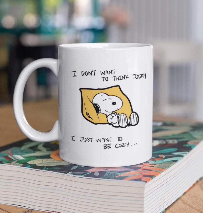 May include: White ceramic mug featuring Snoopy sleeping on a yellow pillow. The mug has the text "I DON'T WANT TO THINK TODAY" and "I JUST WANT TO BE COZY..." printed in black. The mug has a large, curved handle.