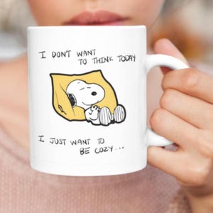 May include: White ceramic mug featuring a Snoopy illustration sleeping on a yellow pillow. The mug has the text "I DON'T WANT TO THINK TODAY, I JUST WANT TO BE COZY..." printed in black. The mug is held by a person.