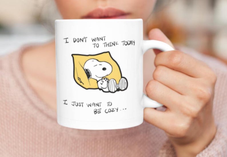 May include: White ceramic mug featuring a Snoopy illustration sleeping on a yellow pillow. The mug has the text "I DON'T WANT TO THINK TODAY, I JUST WANT TO BE COZY..." printed in black. The mug is held by a person.