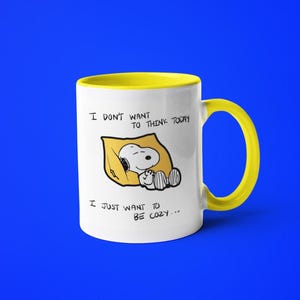 May include: White ceramic mug with a yellow interior and handle. The mug features a cartoon Snoopy character sleeping on a yellow pillow, with the text "I DON'T WANT TO THINK TODAY, I JUST WANT TO BE COZY..." printed on the front.
