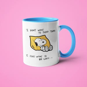 May include: White ceramic mug with a blue interior and handle. The mug features a Snoopy graphic in a yellow blanket with the text "I DON'T WANT TO THINK TODAY, I JUST WANT TO BE COZY..."