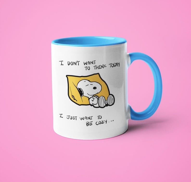 May include: White ceramic mug with a blue interior and handle. The mug features a Snoopy graphic in a yellow blanket with the text "I DON'T WANT TO THINK TODAY, I JUST WANT TO BE COZY..."