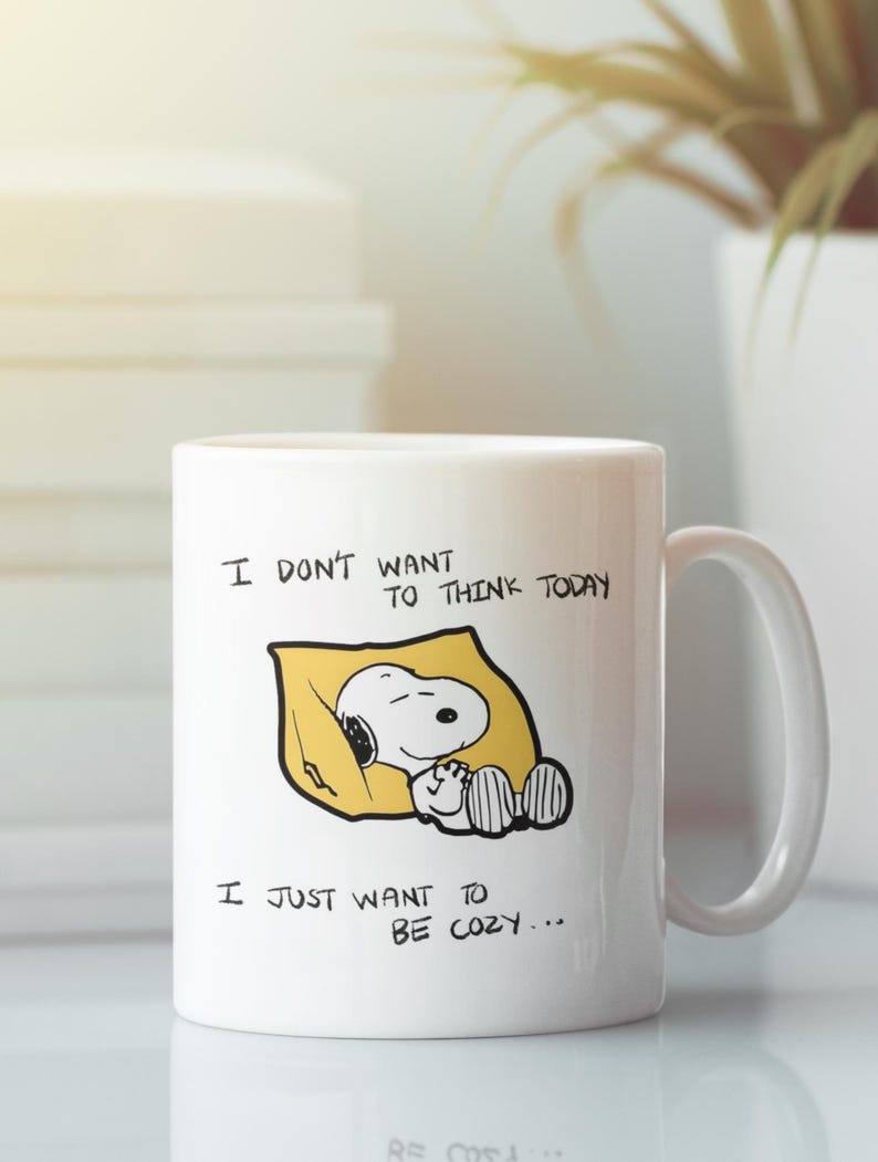 May include: White ceramic mug featuring Snoopy sleeping on a yellow pillow. The mug has the text "I DON'T WANT TO THINK TODAY, I JUST WANT TO BE COZY..." in black lettering. The mug has a white handle and is suitable for hot or cold beverages.