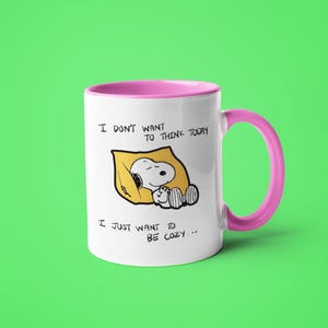 May include: White ceramic mug with a pink interior and handle. The mug features a cartoon Snoopy character sleeping on a yellow pillow. Text on the mug reads "I don't want to think today, I just want to be cozy..."