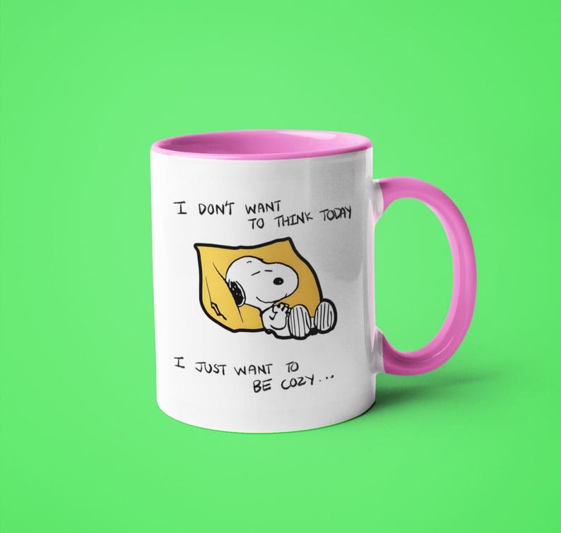 May include: White ceramic mug with a pink interior and handle. The mug features a cartoon Snoopy character sleeping on a yellow pillow. Text on the mug reads "I don't want to think today, I just want to be cozy..."