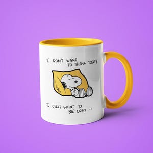 May include: White ceramic mug with a yellow interior and handle. The mug features a Snoopy illustration resting on a yellow pillow, with the text "I DON'T WANT TO THINK TODAY, I JUST WANT TO BE COZY..." in black.
