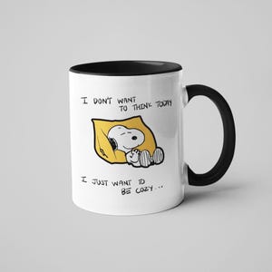 May include: White ceramic mug with a black interior and handle. The mug features a cartoon illustration of Snoopy sleeping in a yellow pillow, with the text "I DON'T WANT TO THINK TODAY, I JUST WANT TO BE COZY..."