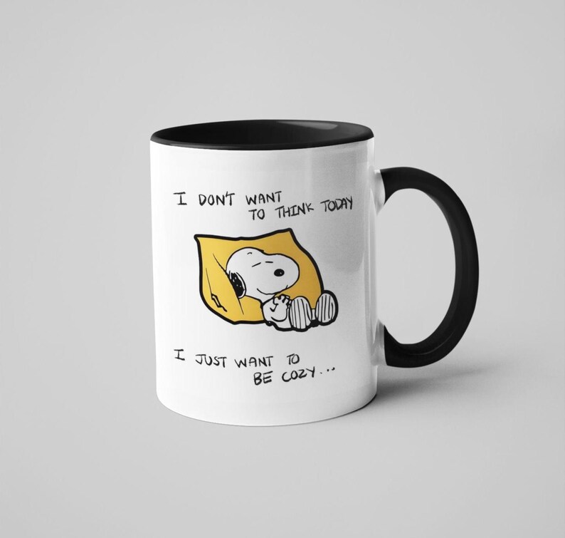 May include: White ceramic mug with a black interior and handle. The mug features a cartoon illustration of Snoopy sleeping in a yellow pillow, with the text "I DON'T WANT TO THINK TODAY, I JUST WANT TO BE COZY..."