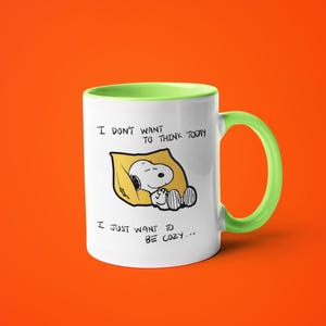 May include: White ceramic mug with a green handle and rim. The mug features a Snoopy illustration lying on a yellow pillow, with the text "I DON'T WANT TO THINK TODAY, I JUST WANT TO BE COZY..."