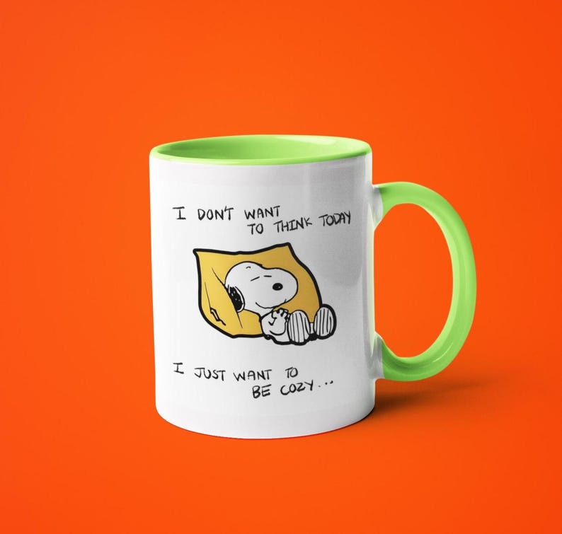 May include: White ceramic mug with a green handle and rim. The mug features a Snoopy illustration lying on a yellow pillow, with the text "I DON'T WANT TO THINK TODAY, I JUST WANT TO BE COZY..."
