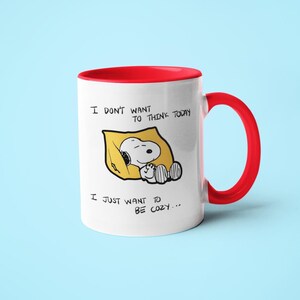 May include: White ceramic mug with a red interior and handle. The mug features a Snoopy illustration in a yellow blanket with the text "I DON'T WANT TO THINK TODAY, I JUST WANT TO BE COZY..."