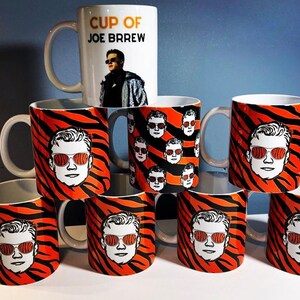 May include: A set of nine white ceramic mugs featuring a tiger stripe pattern and a black and white illustration of a person wearing sunglasses. One mug has the text "Cup of Joe Brew" and an illustration of a person wearing sunglasses.