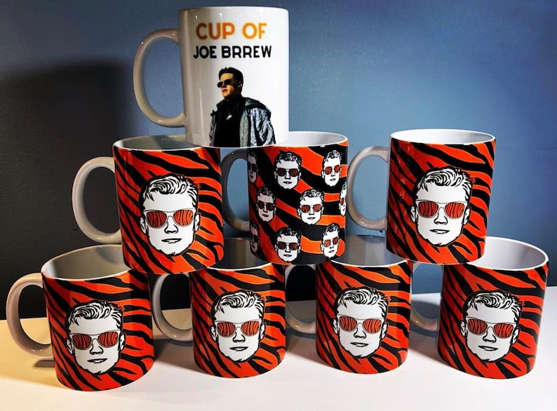 May include: A set of nine white ceramic mugs featuring a tiger stripe pattern and a black and white illustration of a person wearing sunglasses. One mug has the text "Cup of Joe Brew" and an illustration of a person wearing sunglasses.