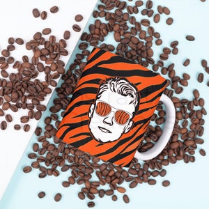 May include: A white ceramic mug with a black and orange tiger stripe pattern. The mug features a graphic of a person wearing sunglasses with a black and orange tiger stripe pattern.