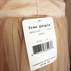 May include: A close-up of a beige skirt with a tulle overlay. The skirt has a satin waistband and a tag that reads "free people", size 12, and "SHELL". The suggested retail price is $148.00.