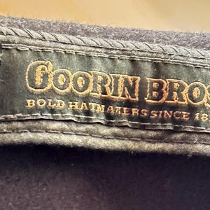 May include: A dark charcoal gray hat featuring an interior black fabric label with gold embroidered text that reads Goorin Bros. Bold Hatmakers Since 1895. The label is stitched onto the inner band of the hat with visible dark thread detailing.