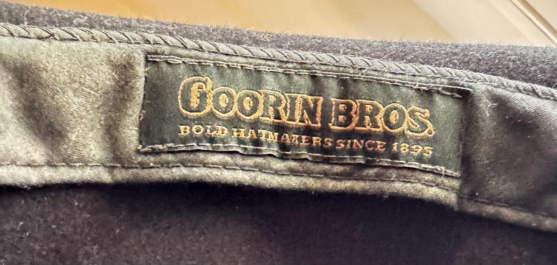 May include: A dark charcoal gray hat featuring an interior black fabric label with gold embroidered text that reads Goorin Bros. Bold Hatmakers Since 1895. The label is stitched onto the inner band of the hat with visible dark thread detailing.