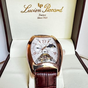 May include: A gold-toned Lucien Piccard automatic watch with a white face, visible gears, and a brown leather band. The watch is displayed in a cream-colored box with the brand name and "Swiss Since 1923" printed on the lid.