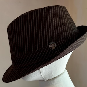 K&ouml;nnte beinhalten: Ein dunkelbrauner Fedora-Hut mit einem dezenten vertikalen Nadelstreifenmuster auf der Krone und der Krempe. Ein kleines, metallisches schildf&ouml;rmiges Emblem mit der Aufschrift "Lester Young" ist seitlich direkt &uuml;ber der Krempe angebracht.