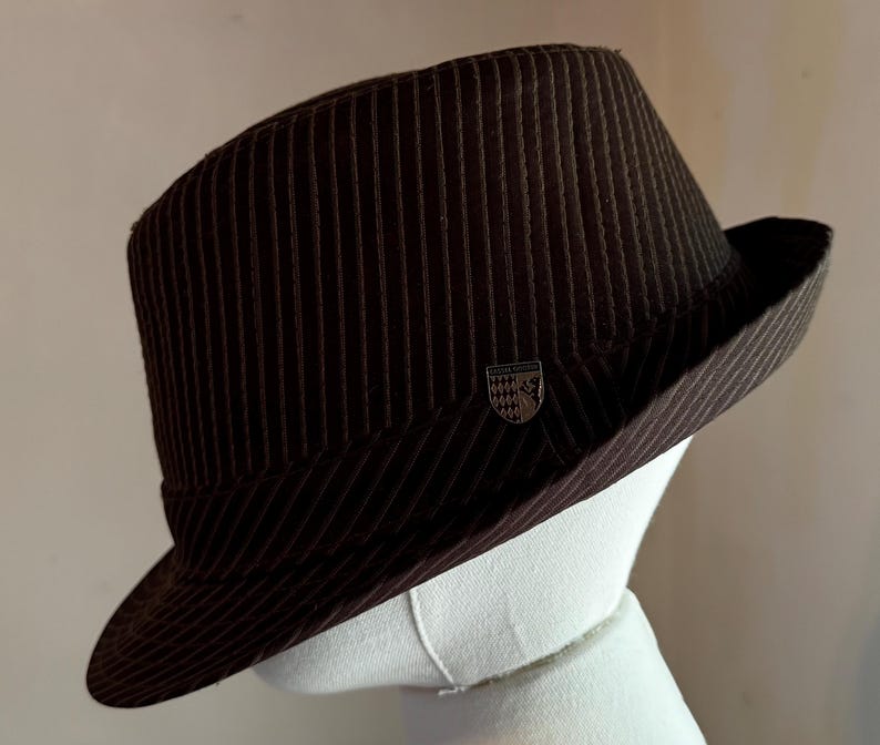 K&ouml;nnte beinhalten: Ein dunkelbrauner Fedora-Hut mit einem dezenten vertikalen Nadelstreifenmuster auf der Krone und der Krempe. Ein kleines, metallisches schildf&ouml;rmiges Emblem mit der Aufschrift "Lester Young" ist seitlich direkt &uuml;ber der Krempe angebracht.