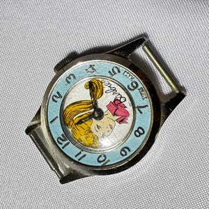 May include: A round silver-tone wristwatch featuring a light blue bezel with black numerals from 1 to 12. The watch face displays a graphic illustration of a blonde character with the text Barbie printed in black script near the center.