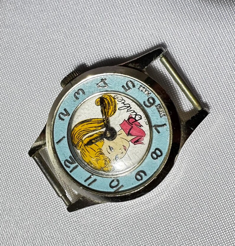 May include: A round silver-tone wristwatch featuring a light blue bezel with black numerals from 1 to 12. The watch face displays a graphic illustration of a blonde character with the text Barbie printed in black script near the center.
