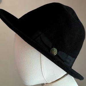 May include: A black felt fedora hat featuring a wide brim and a black grosgrain ribbon band. The hat is accented with a circular metallic button on the side, which displays an embossed crown emblem centered on the dark ribbon detail.