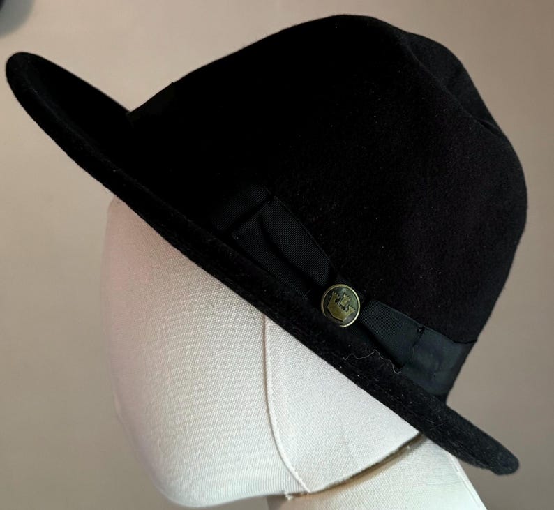 May include: A black felt fedora hat featuring a wide brim and a black grosgrain ribbon band. The hat is accented with a circular metallic button on the side, which displays an embossed crown emblem centered on the dark ribbon detail.