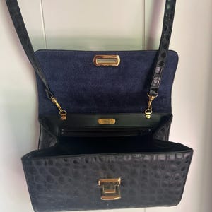 May include: Black crocodile embossed leather crossbody bag with a gold clasp and a blue suede lining. The bag has a small pocket on the inside and a long strap.