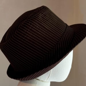 K&ouml;nnte beinhalten: Ein dunkelbrauner Fedora-Hut mit einem dezenten vertikalen Nadelstreifenmuster auf der Krone und der Krempe. Der Hut hat eine strukturierte Form mit einer eingekniffenen Krone und einer leicht nach oben gebogenen Krempe in dunklem Farbton.