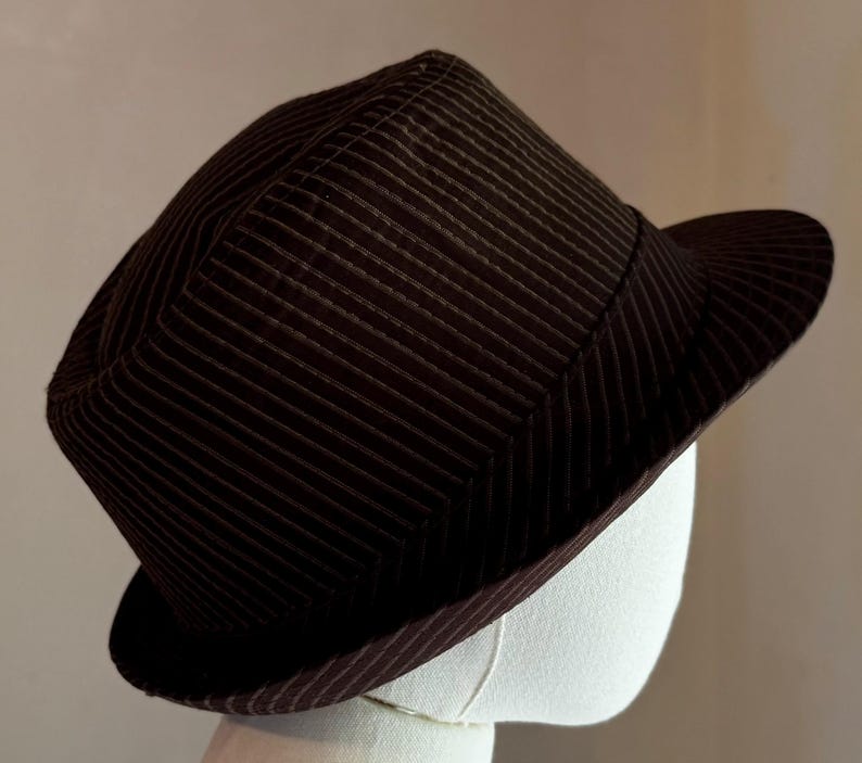 K&ouml;nnte beinhalten: Ein dunkelbrauner Fedora-Hut mit einem dezenten vertikalen Nadelstreifenmuster auf der Krone und der Krempe. Der Hut hat eine strukturierte Form mit einer eingekniffenen Krone und einer leicht nach oben gebogenen Krempe in dunklem Farbton.