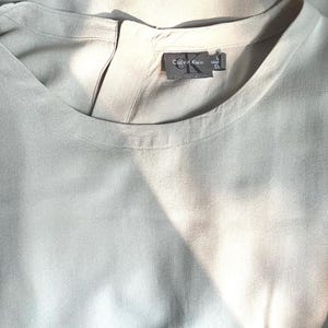 Könnte beinhalten: Eine hellgraue Calvin Klein Bluse. Das Oberteil hat einen runden Ausschnitt und einen kleinen Schlitz am Rücken. Das Calvin Klein-Etikett ist in der Nähe des Ausschnitts sichtbar. Der Stoff scheint ein weiches, fließendes Material zu sein.