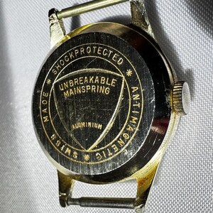 May include: A gold-tone watch case back with a black circular center featuring the text SHOCKPROTECTED, ANTIMAGNETIC, SWISS MADE, UNBREAKABLE MAINSPRING, and ALUMINIUM. The metal casing has a metallic finish and includes two spring bars.