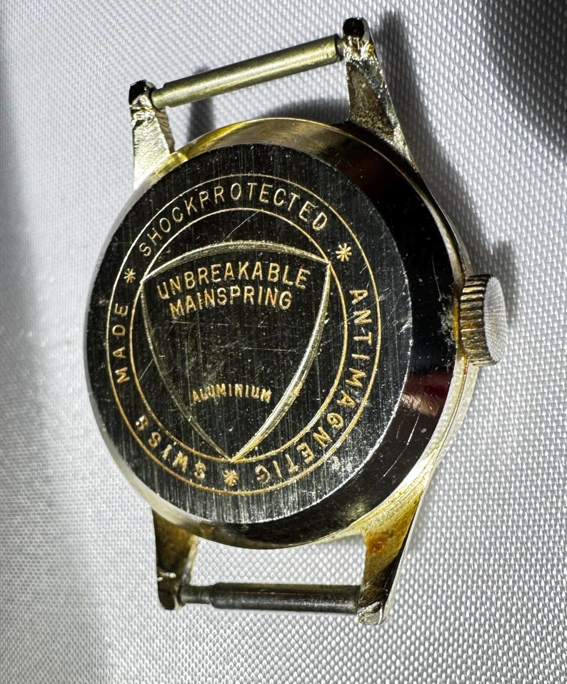 May include: A gold-tone watch case back with a black circular center featuring the text SHOCKPROTECTED, ANTIMAGNETIC, SWISS MADE, UNBREAKABLE MAINSPRING, and ALUMINIUM. The metal casing has a metallic finish and includes two spring bars.