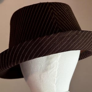 K&ouml;nnte beinhalten: Ein dunkelbrauner Fedora-Hut mit einem dezenten diagonalen Nadelstreifenmuster auf der Krone und der Krempe. Der Hut wird auf einem wei&szlig;en Schaufensterpuppenkopf pr&auml;sentiert und zeigt die strukturierte Form sowie die geschwungene Krempe.