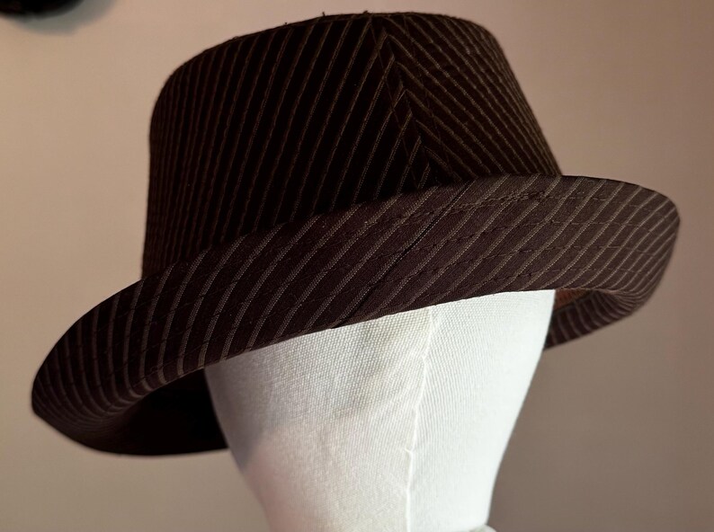 K&ouml;nnte beinhalten: Ein dunkelbrauner Fedora-Hut mit einem dezenten diagonalen Nadelstreifenmuster auf der Krone und der Krempe. Der Hut wird auf einem wei&szlig;en Schaufensterpuppenkopf pr&auml;sentiert und zeigt die strukturierte Form sowie die geschwungene Krempe.