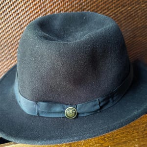 May include: A black felt fedora hat featuring a structured crown and a wide brim. The hat is accented with a black grosgrain ribbon band that encircles the base of the crown, finished with a small, circular gold-toned metal emblem at the front.