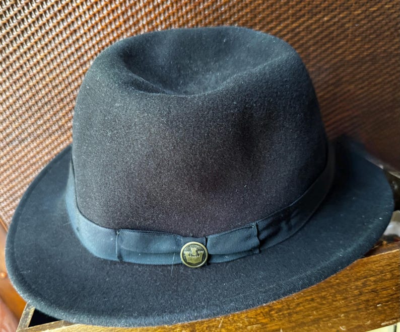 May include: A black felt fedora hat featuring a structured crown and a wide brim. The hat is accented with a black grosgrain ribbon band that encircles the base of the crown, finished with a small, circular gold-toned metal emblem at the front.