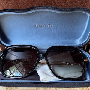 May include: Black sunglasses with a square frame and dark lenses, resting in a blue velvet case. The case is open, revealing the Gucci logo in gold lettering. A white cleaning cloth is also inside the case.